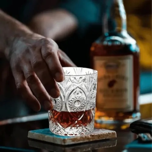 Mana unui barman care ridica un pahar sculptat plin cu whiskey de pe un suport de piatra, cu o sticla de alcool in fundal.
