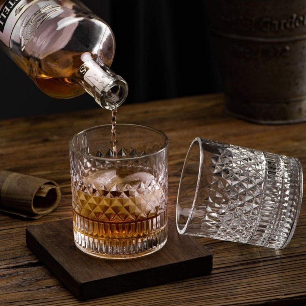 pahare de whisky din cristal masiv MADGENIX Noble pline cu bourbon si cuburi de gheata, asezate pe o masa intunecata de bar, cu o atmosfera eleganta.