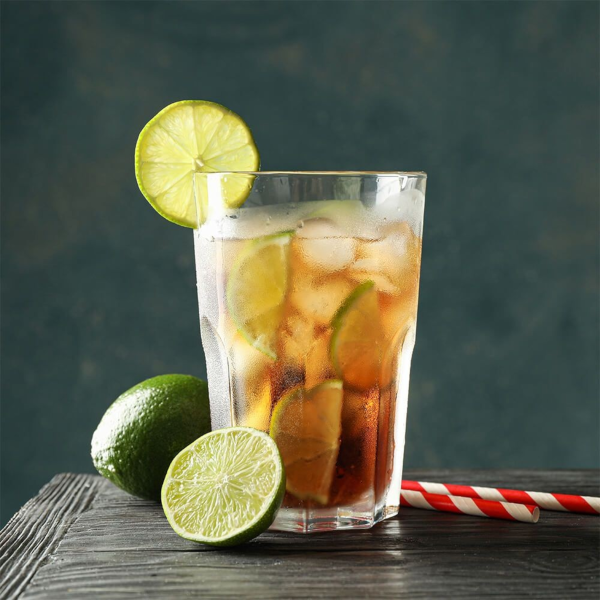 Pahar Highball plin cu ice tea, cuburi de gheata si felii de lime, asezat pe o masa intunecata langa lime proaspat.